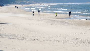 Una playa con gente caminando por la orilla, cielo despejado y un área boscosa al fondo.