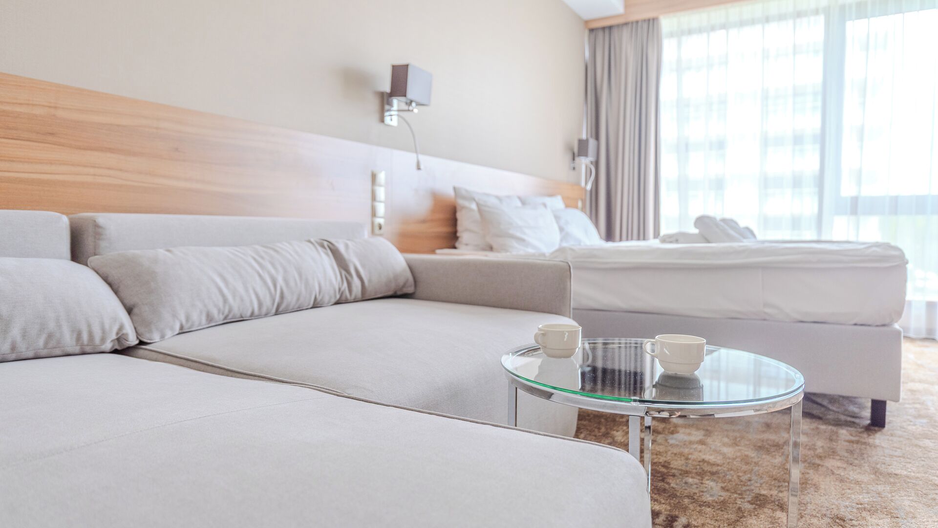 A modern hotel room with a large sofa, a smaller bed, a glass coffee table with two cups, and a large window with curtains.