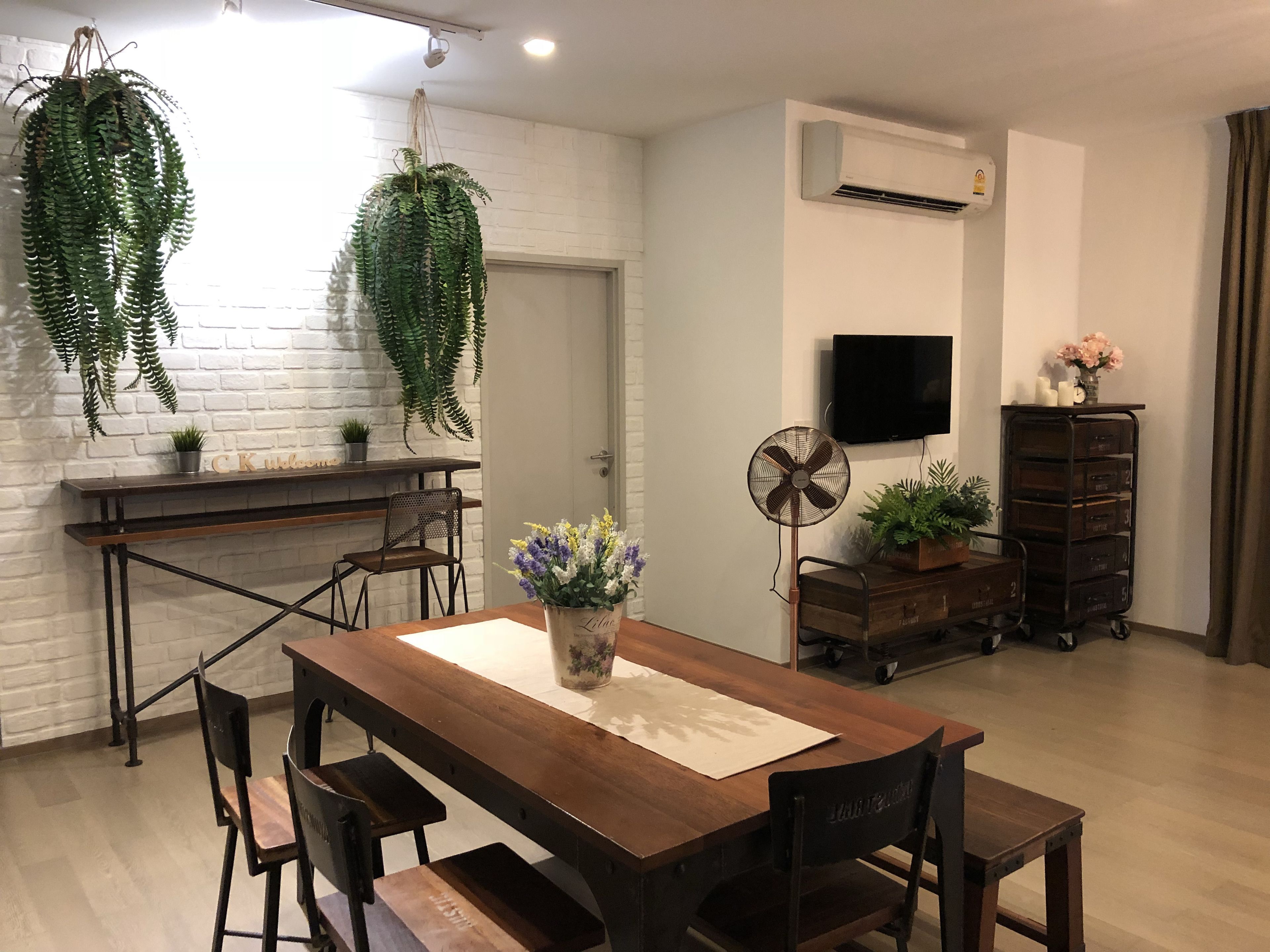 Un comedor con una mesa de madera, sillas, plantas colgantes, un ventilador, un televisor y un mueble de almacenamiento con cajones.