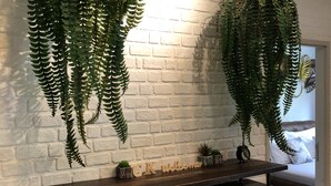 Una habitación con pared de ladrillo blanco, una mesa de madera con un cartel de 'Welcome', dos helechos colgantes, una silla de metal y una cama al fondo.