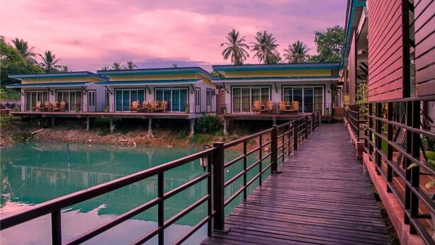 A wooden bridge leading to a row of bungalow-style buildings with large windows and a view of a calm body of water. - Laketerrace Resort (La-ngu)