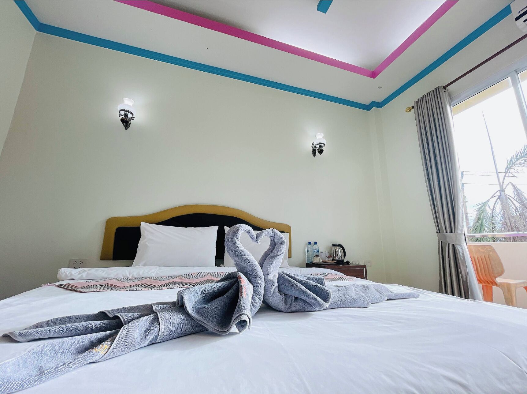 Una habitación de hotel con cama, mesita de noche, ventana con cortinas y vista a palmeras.