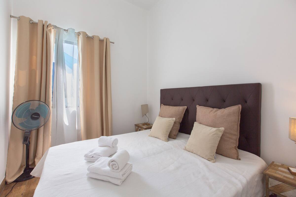 A neatly made bed with white linens and rolled towels, a dark upholstered headboard, beige curtains, and a standing fan.