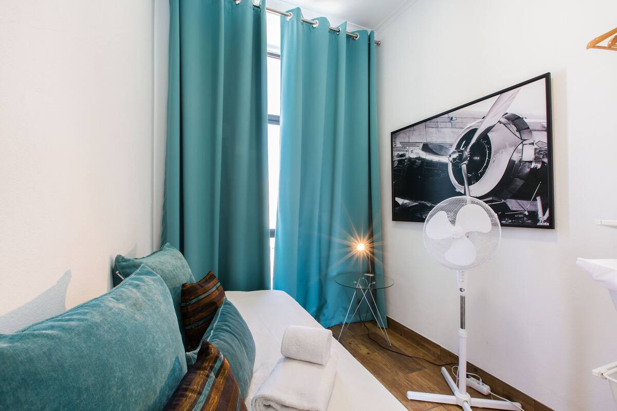 A hotel room with teal curtains, a bed with a white towel, a standing fan, and a framed picture on the wall.