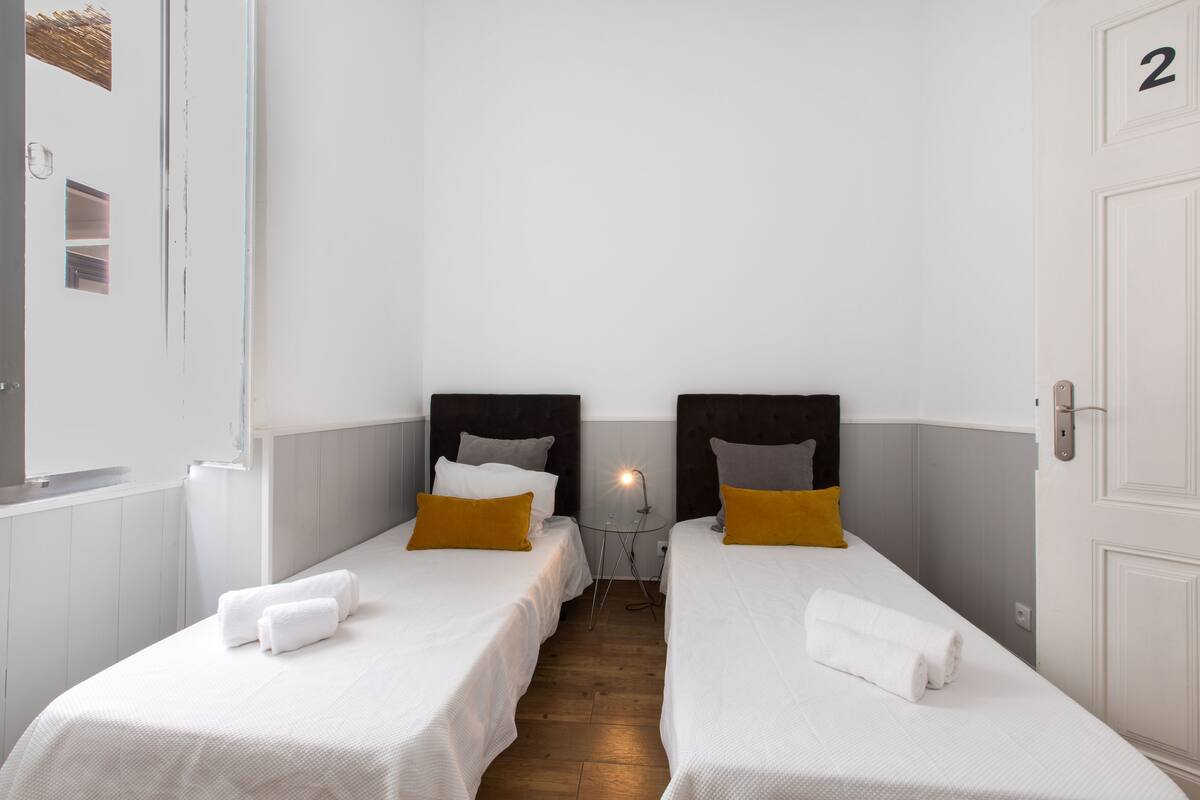 A hotel room with two single beds, each with a white bedspread and a single pillow. There is a small table with a lamp between the beds, and a white door with the number '2' above it.