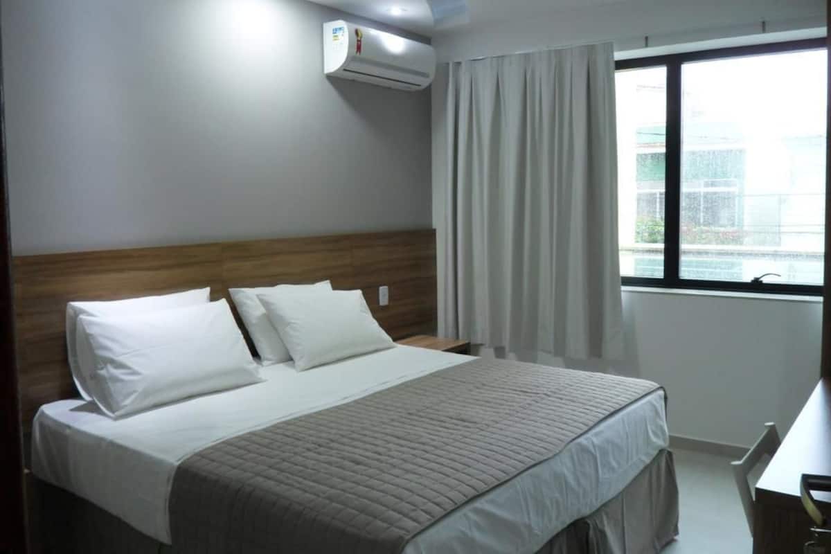 Habitación de hotel con cama, cabecera, ventana con cortinas, equipo de aire acondicionado y escritorio con silla.