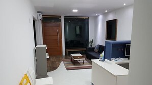 Una sala de estar moderna con un sofá, una mesa de centro y un televisor. Se observa una puerta de madera y una ventana con persianas.