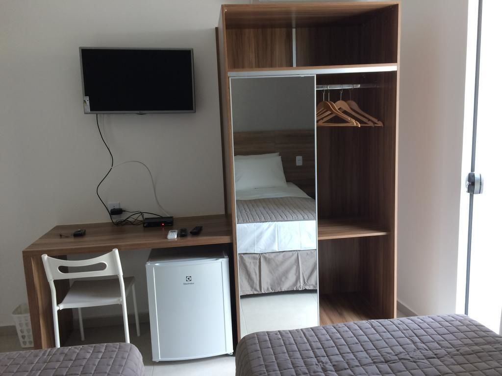 A compact room with a bed, a desk, a small fridge, a TV mounted on the wall, and a wardrobe with hanging space.