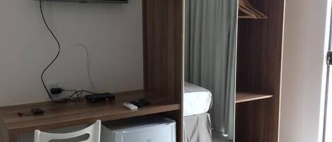 A hotel room with a bed, a desk with a chair, a small refrigerator, a wardrobe, and a television mounted on the wall.
