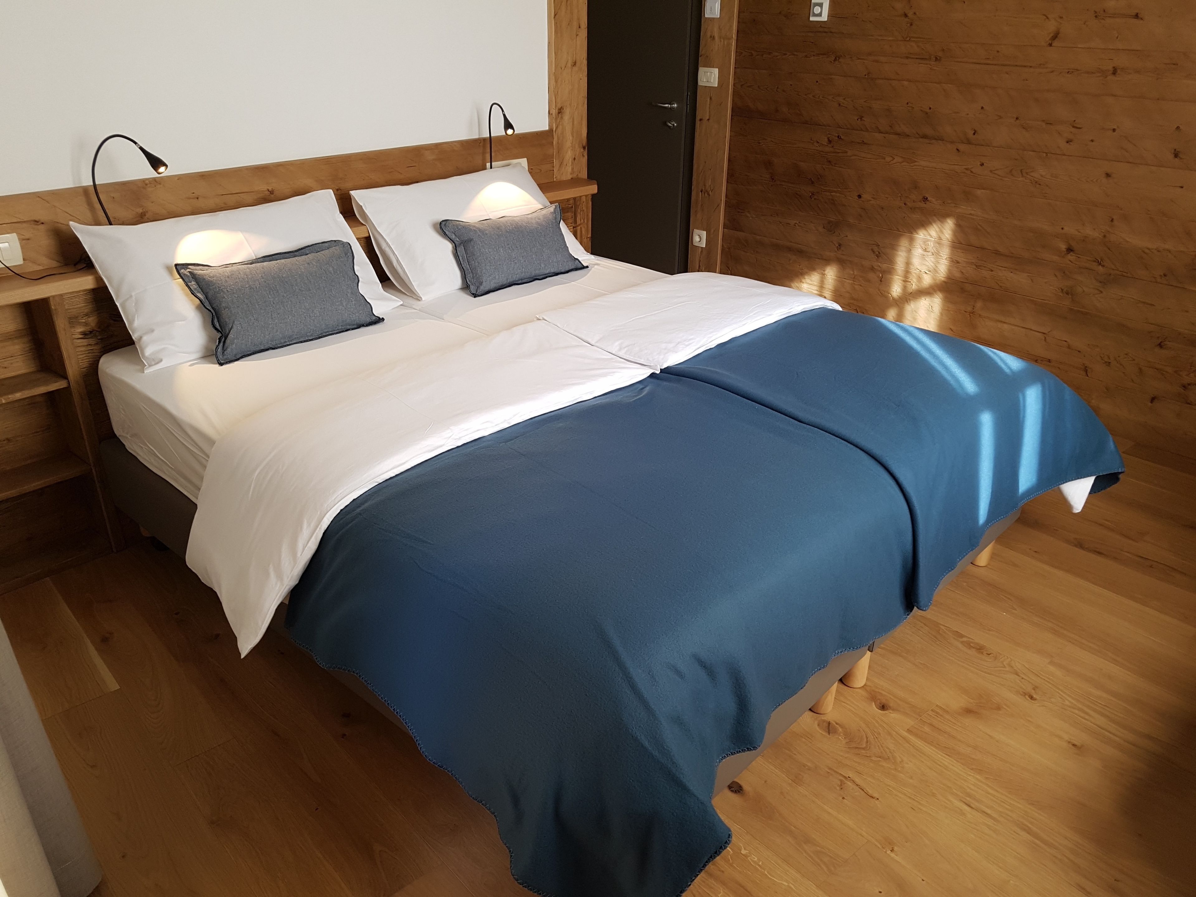 Una cama con ropa de cama blanca y azul, cabecera de madera, lámparas de noche y suelo de madera.
