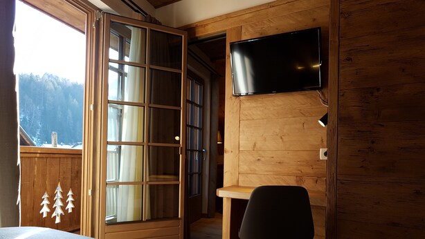 Una habitación con cama, escritorio, silla, televisión y un amplio ventanal con cortinas.