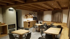 Un comedor moderno con mesas y sillas de madera, una zona de cocina con cafetera y una sala de estar con cortinas.