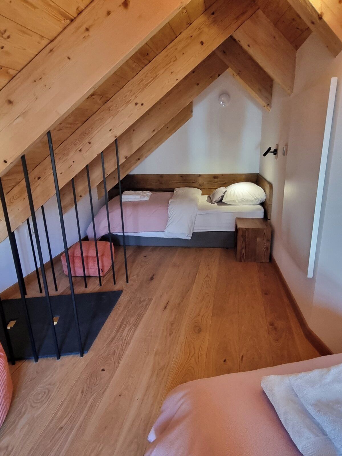Une chambre avec un plafond en pente, des poutres en bois, un lit, un petit tabouret et un tapis noir.
