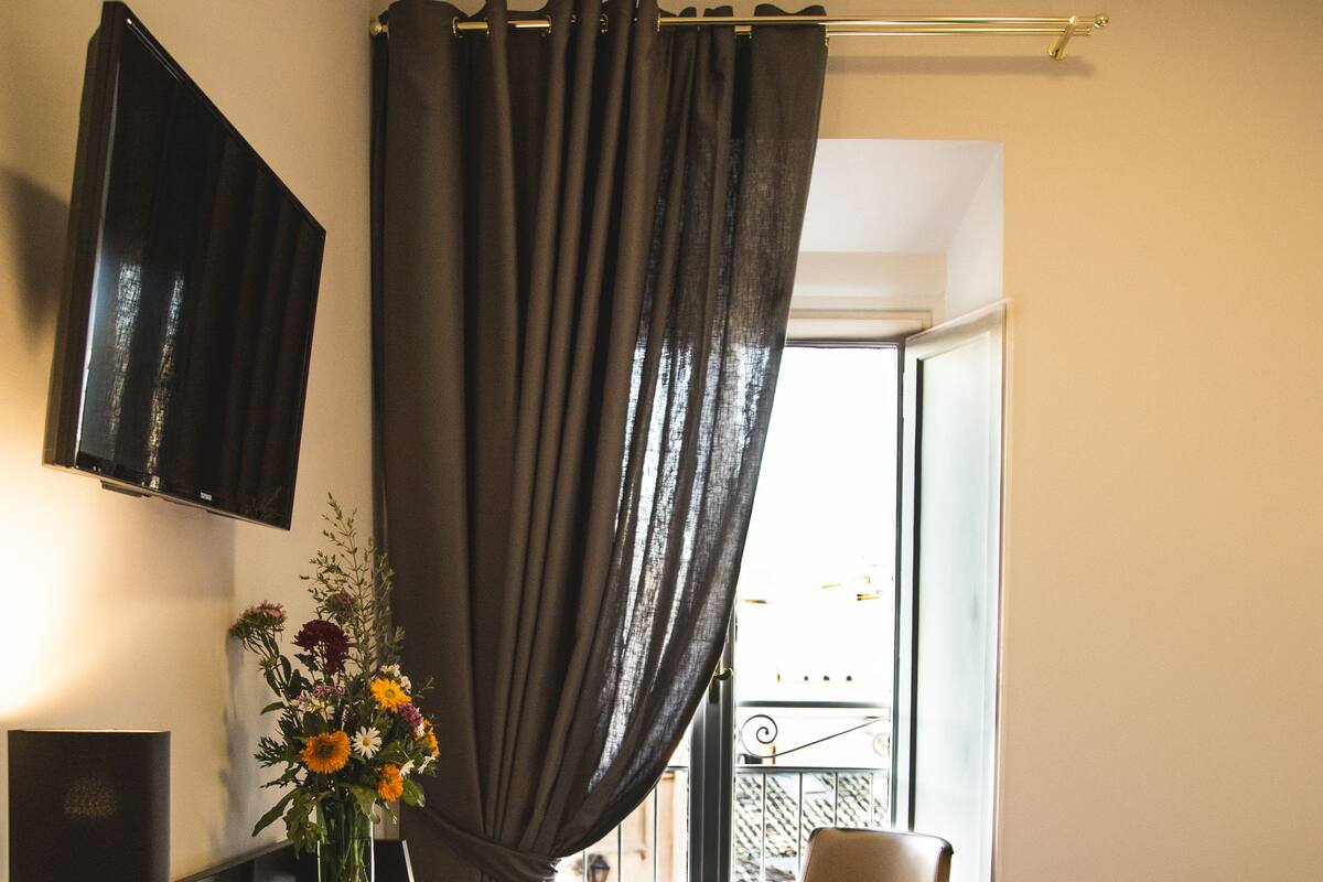 A hotel room with a bed, a desk with a lamp, a chair, a TV mounted on the wall, and a window with curtains.