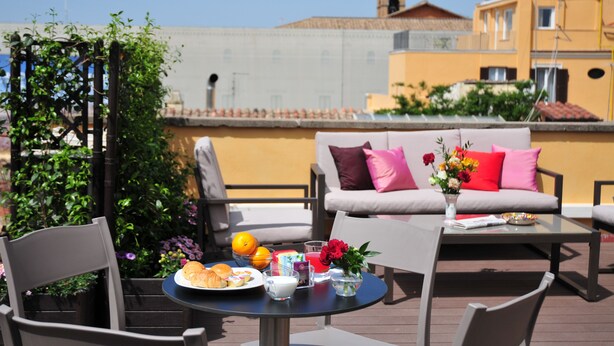 A rooftop terrace with outdoor seating, a table with breakfast items, and a view of buildings in the background. - Piccola Navona Roof Garden (Rome)