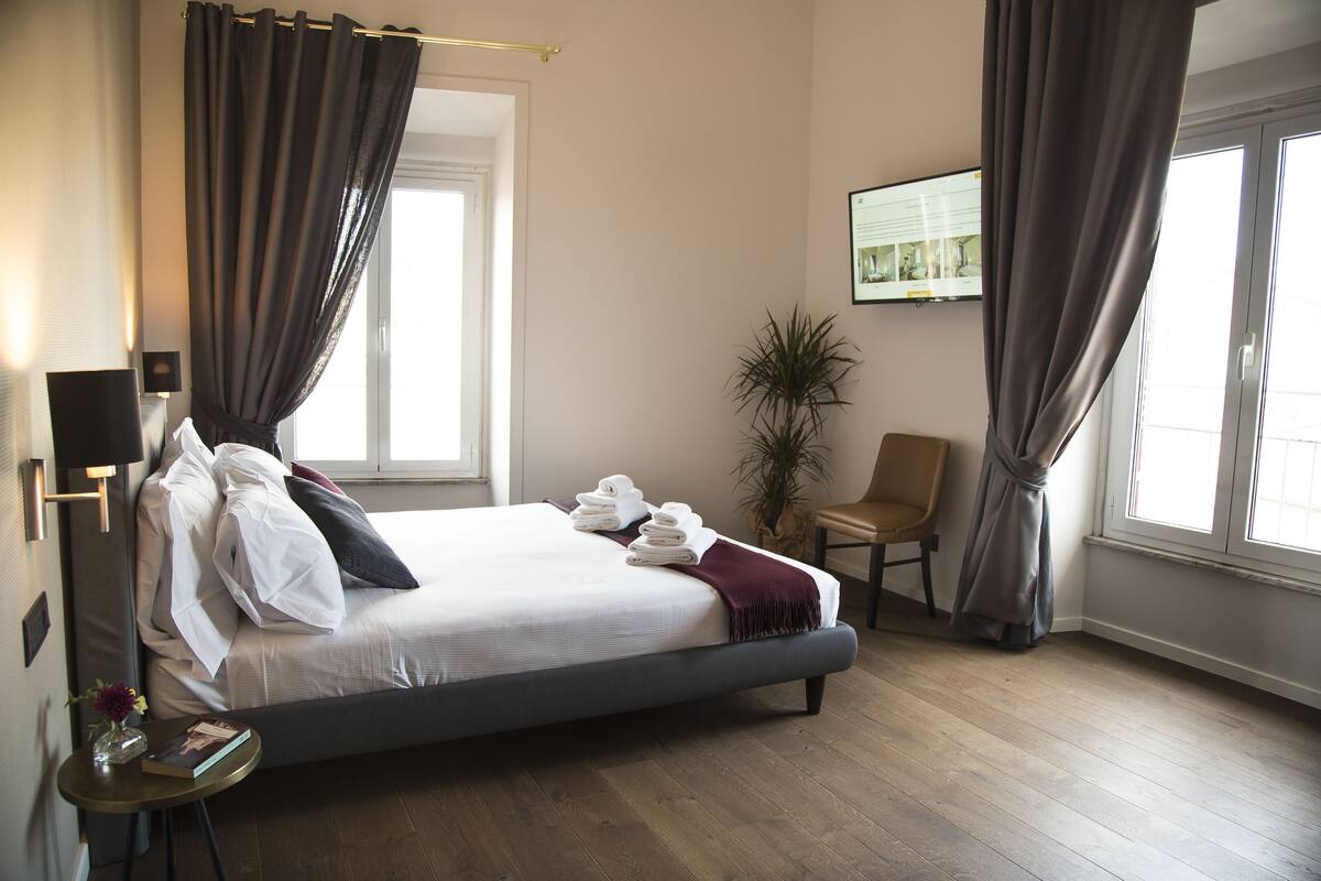 A neatly made bed with white linens and a dark headboard, a small round table with a book and vase, a chair, and a potted plant in a modern hotel room with wooden flooring and large windows.