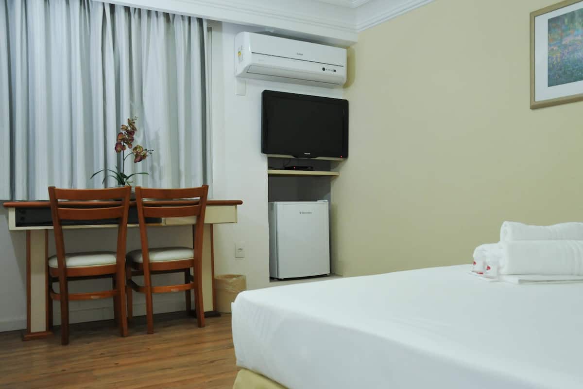 Ein Hotelzimmer mit einem Bett, einem Schreibtisch mit zwei Stühlen, einem Fernseher, einem kleinen Kühlschrank und einem Fenster mit Vorhängen.