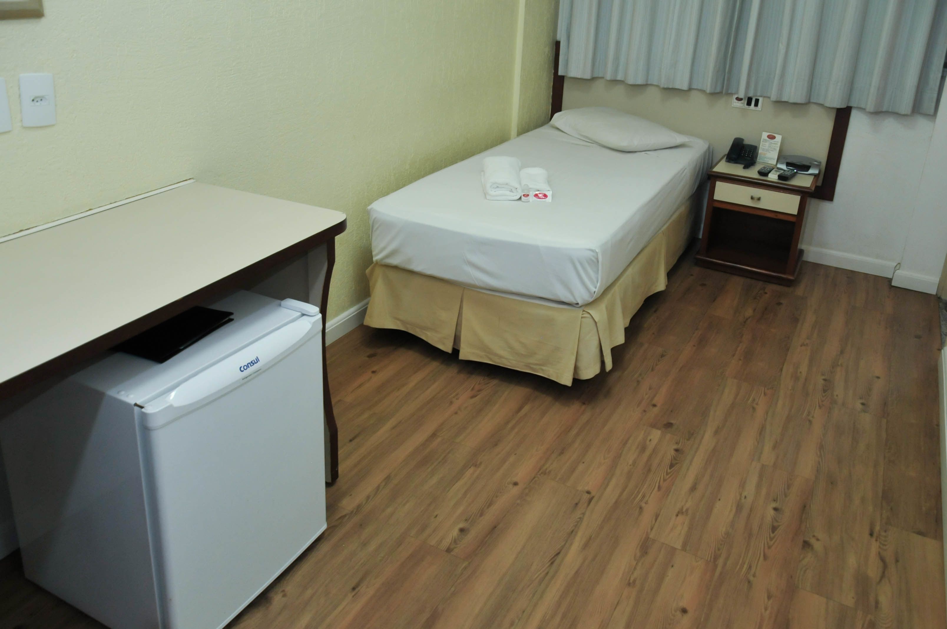 Una habitación pequeña y limpia con una cama, una mesita de noche con teléfono y lámpara, un escritorio con refrigerador y una ventana con cortinas.