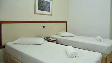 Habitación de hotel con dos camas individuales, cada una con una almohada y una toalla enrollada. Se ve una mesita de noche con un teléfono y un objeto pequeño encima. Encima de las camas, hay un cuadro enmarcado colgado en la pared.