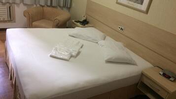 Ein ordentlich bezogenes Bett mit weißen Leinen und Kissen, ein Nachttisch mit Telefon und Uhr, ein Sessel und ein Bild an der Wand.