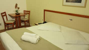 Una cama bien hecha con ropa de cama blanca, un edredón verde y toallas enrolladas. Hay una mesita con un florero y dos sillas. Un cuadro enmarcado cuelga de la pared sobre la cama.