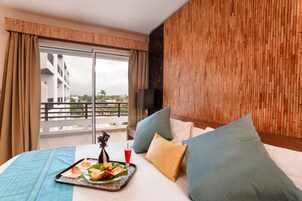 Habitación de hotel con balcón, cama con almohadas, bandeja con comida y bebida, y televisor en la pared.