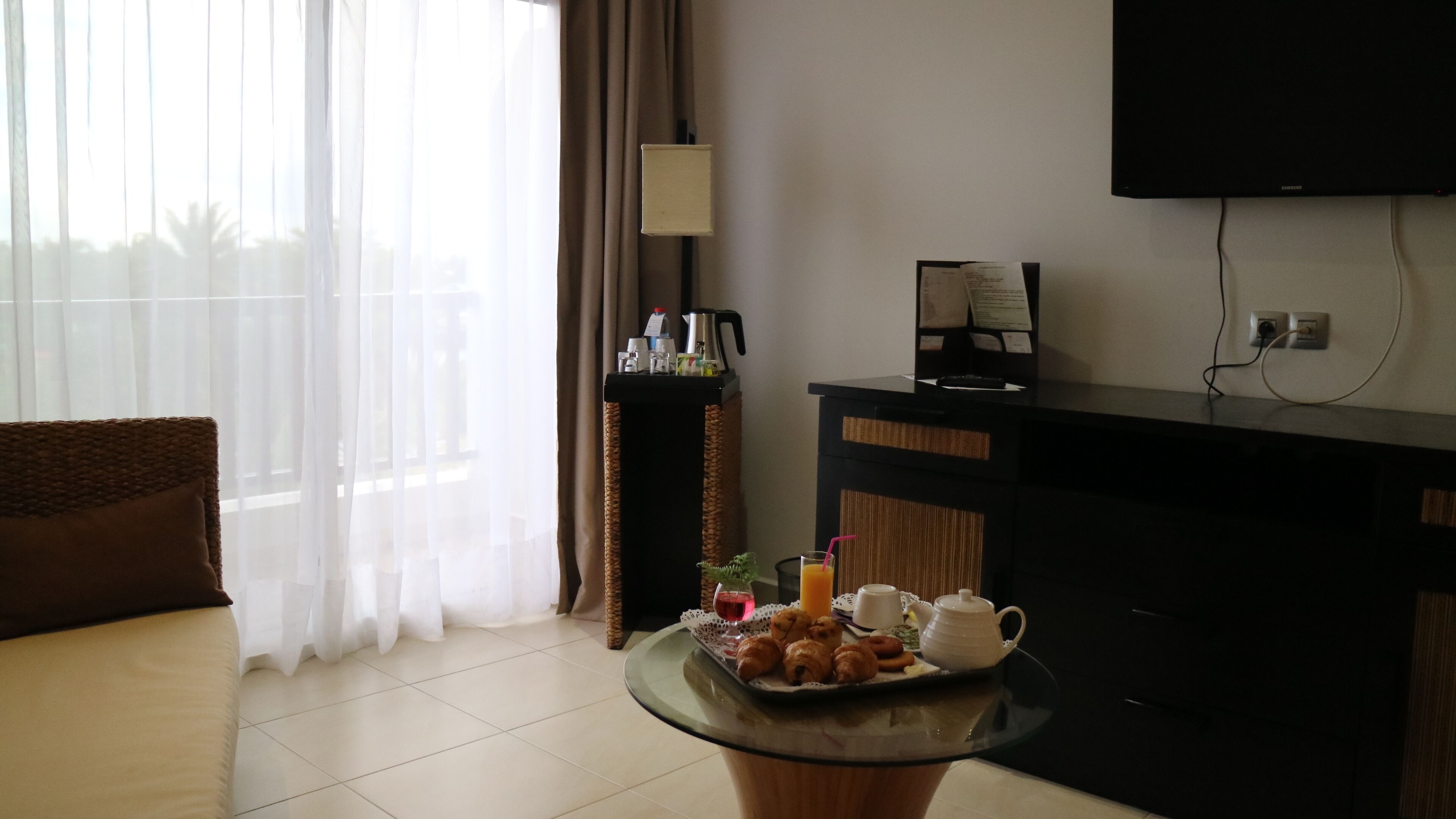 Habitación de hotel con una mesa de comedor dispuesta con cruasanes, un hervidor de té y un vaso de jugo de naranja. Hay un sofá y un televisor en la pared.