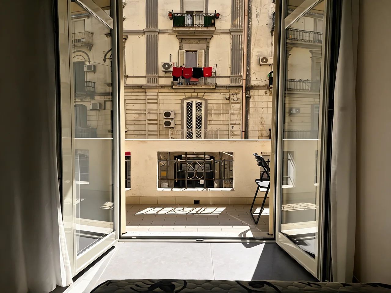 Vista através de uma porta de vidro para um edifício com varanda, uma janela com grade de metal e uma pequena mesa com uma cadeira.