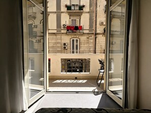 A view through a glass door into a building with a balcony, a window with a metal grille, and a small table with a chair. - Donn'Anna Bed & Breakfast (Naples)