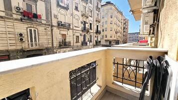 Uma varanda com vista para edifícios próximos, grade de metal e janela com veneziana metálica.
