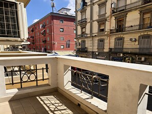 Un balcon doté d’une rambarde en métal, d’un sol carrelé et offrant une vue sur les bâtiments voisins.