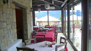 Un área de comedor al aire libre con muros de piedra y ventanas amplias que ofrecen vistas a la montaña. Las mesas están dispuestas con manteles rojos y blancos, y hay sillas de madera.
