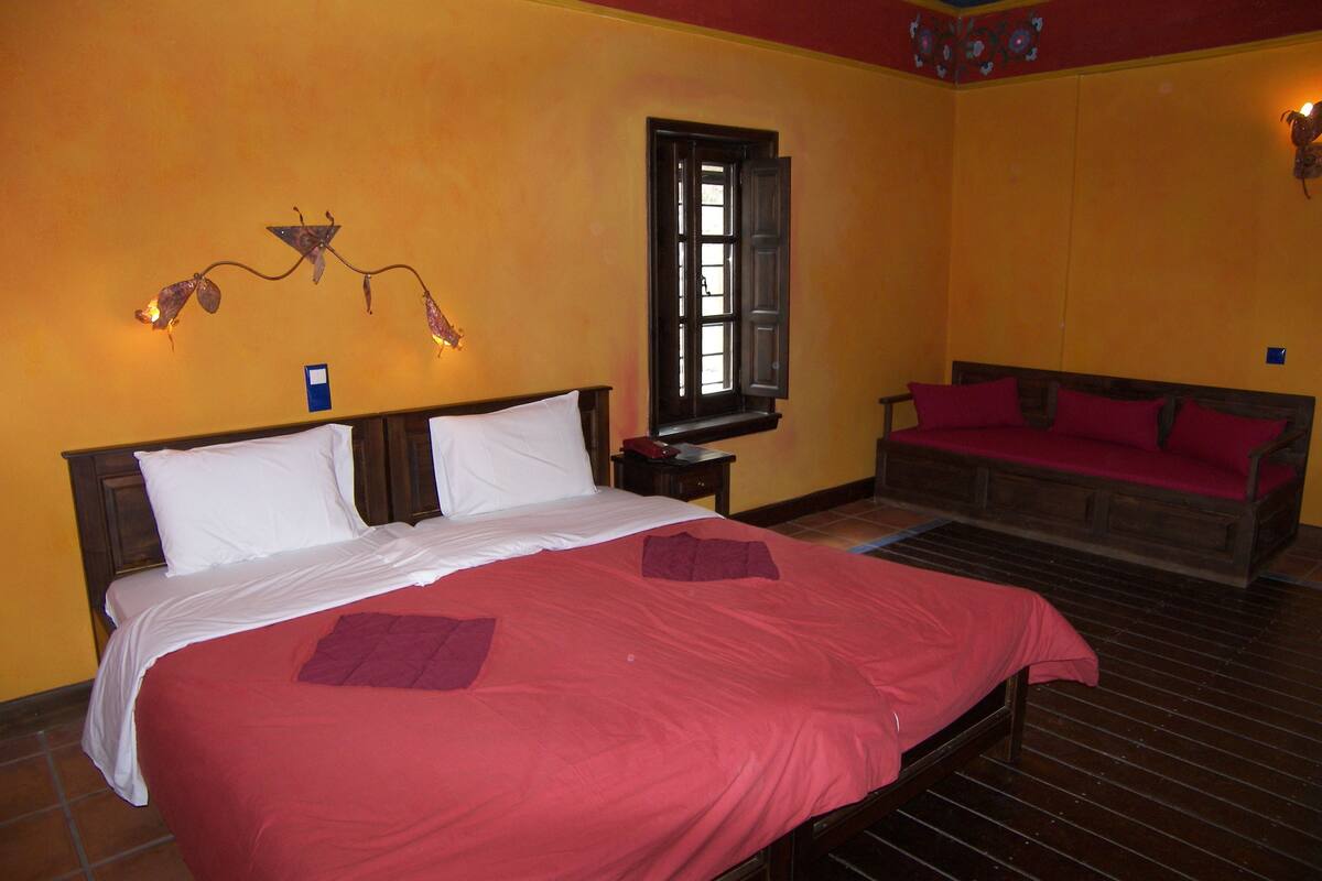 Una cama doble con colcha roja y dos almohadas, una mesita de noche con lámpara, una ventana con persiana de madera y un banco de madera con cojines rojos en una habitación con paredes amarillas.