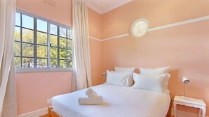 A bedroom with a large window, a bed with white linens, a bedside table with a lamp, and a wall clock. - Kenwood 7 (Cape Town)