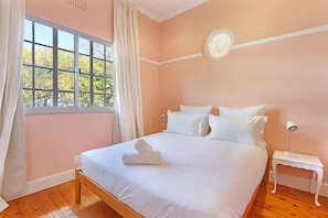 A bedroom with a large window, a bed with white linens, a bedside table with a lamp, and a wall clock.