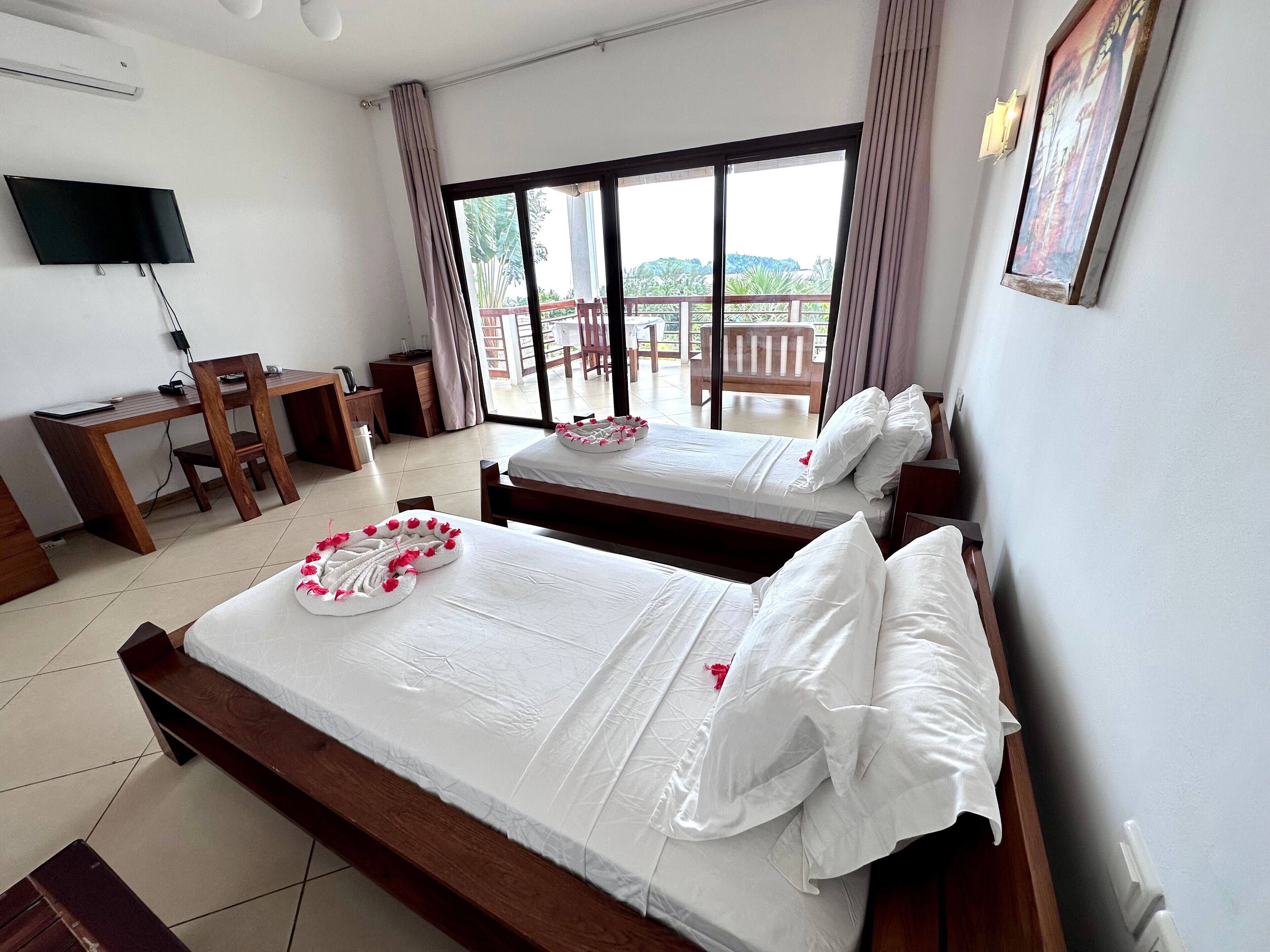 A hotel room with two beds, a desk, a chair, a TV, and a balcony with a view.