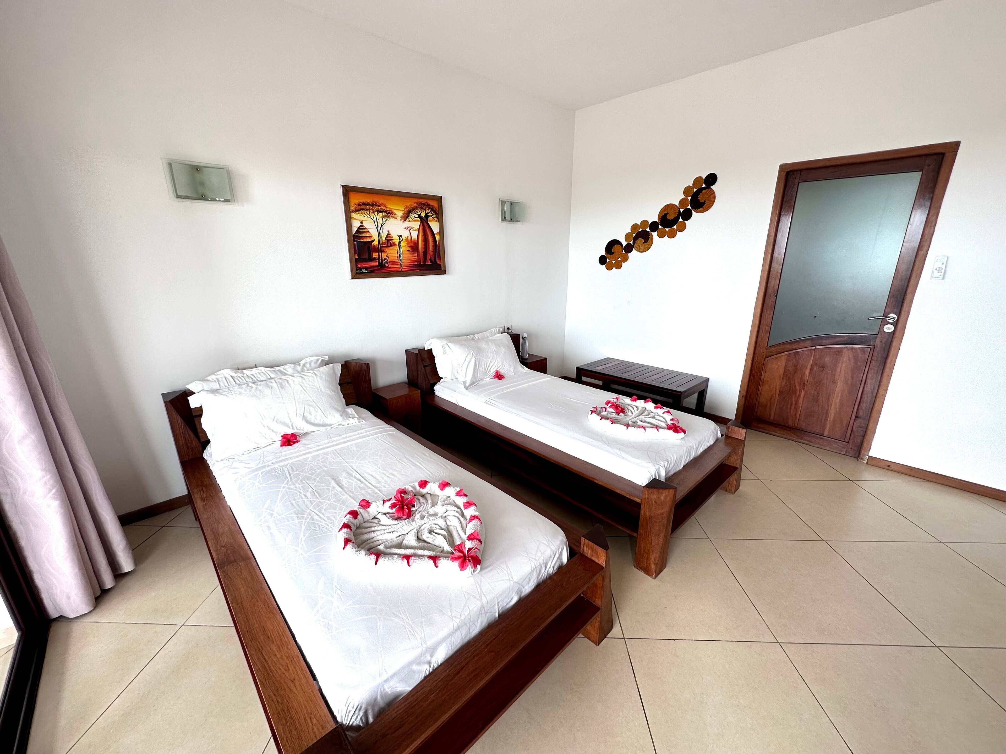 A hotel room with two wooden beds, white bedding, a painting on the wall, a decorative wall piece, and a wooden door.