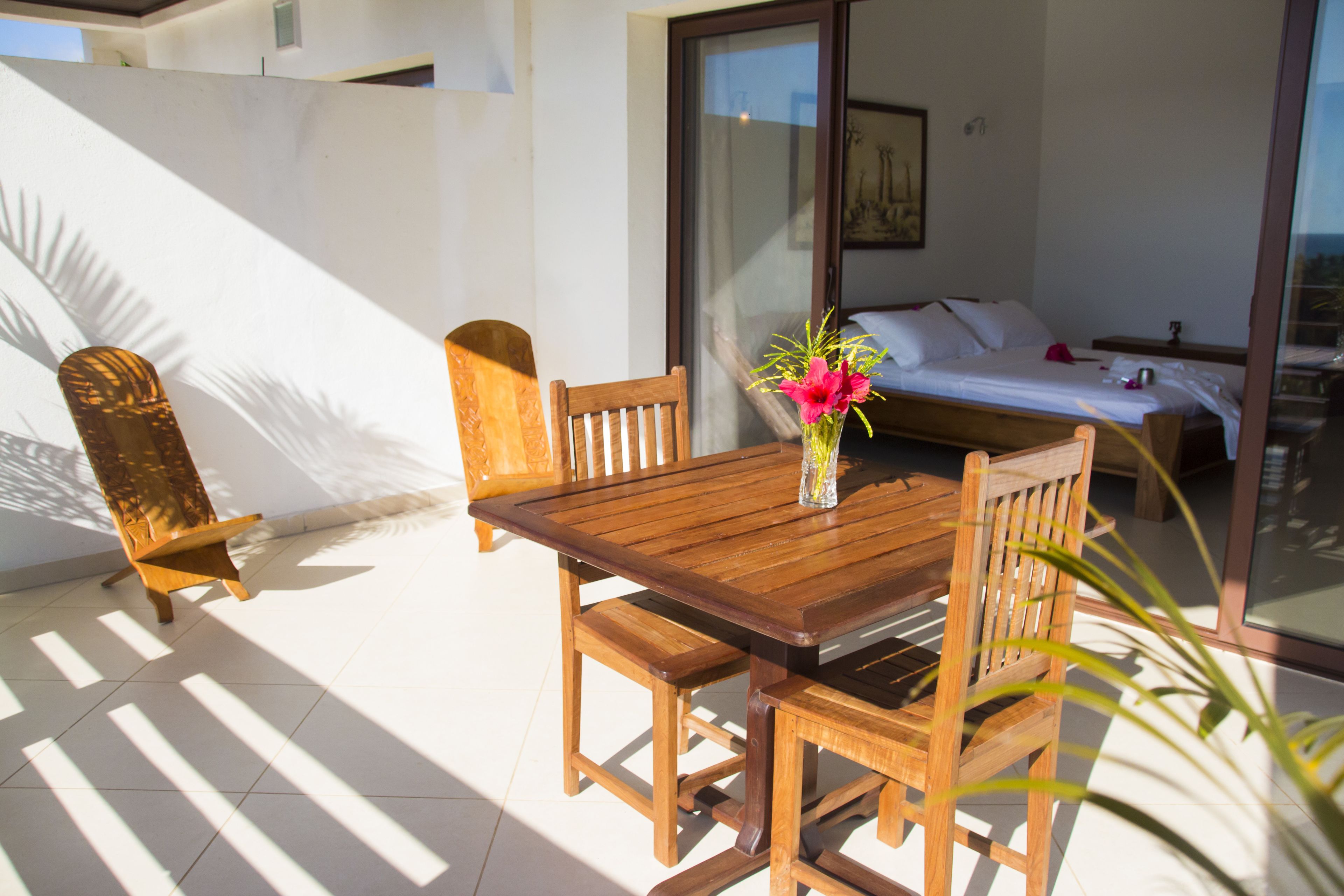 Una mesa de comedor de madera con sillas, un jarrón con flores y una cama con una ventana al fondo.