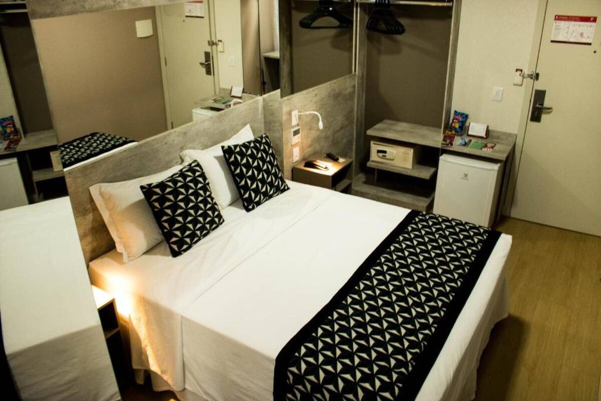 A hotel room with a bed, a nightstand, a mirror, a wardrobe, and a small desk with a lamp.
