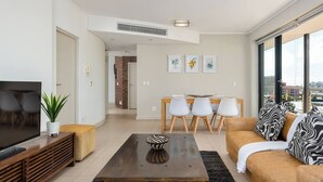 Ein modernes Wohnzimmer mit einem Flachbildfernseher, einem Holztisch und einer Couch mit gemusterten Kissen. Es gibt einen Essbereich mit Tisch und Stühlen sowie ein großes Fenster mit Blick auf die Stadt.