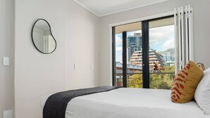 Ein Schlafzimmer mit einem großen Bett, Blick auf Gebäude durch ein Fenster und einem Spiegel an der Wand.