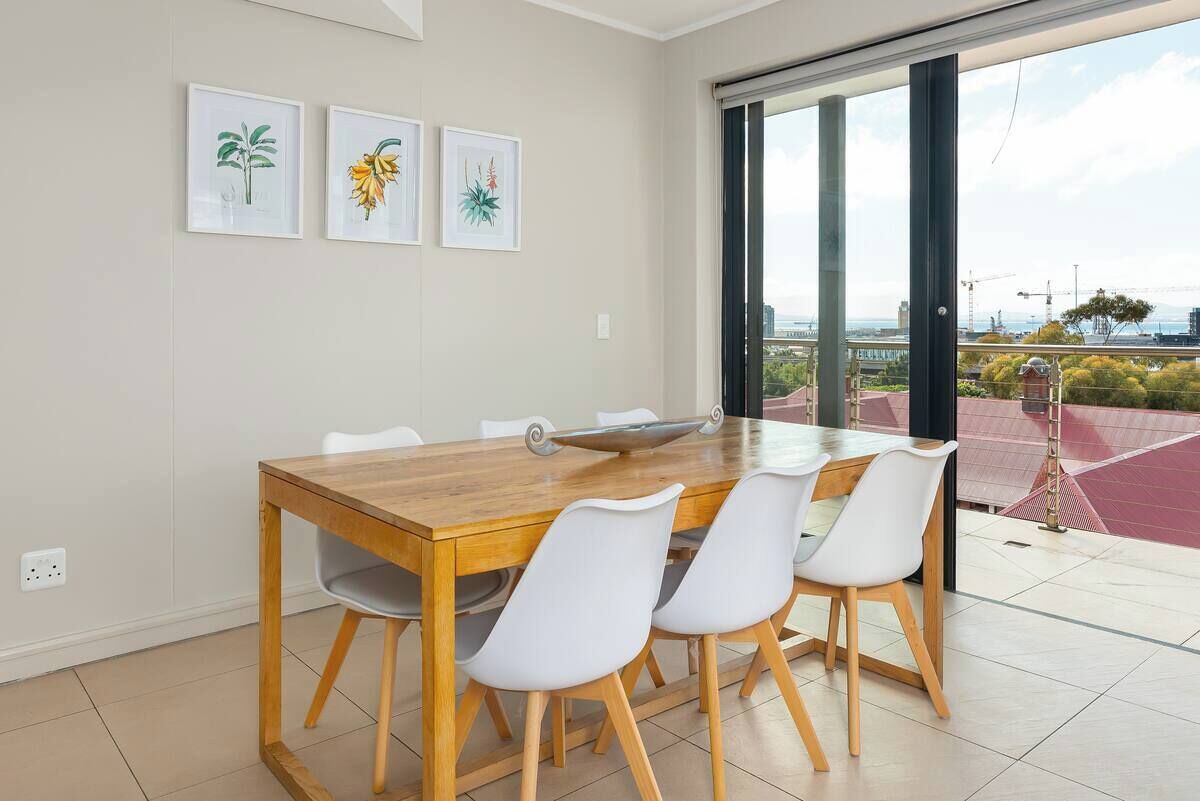 A dining area with a wooden table and white chairs, a large window with a view of a bridge and greenery, and three framed botanical prints on the wall.