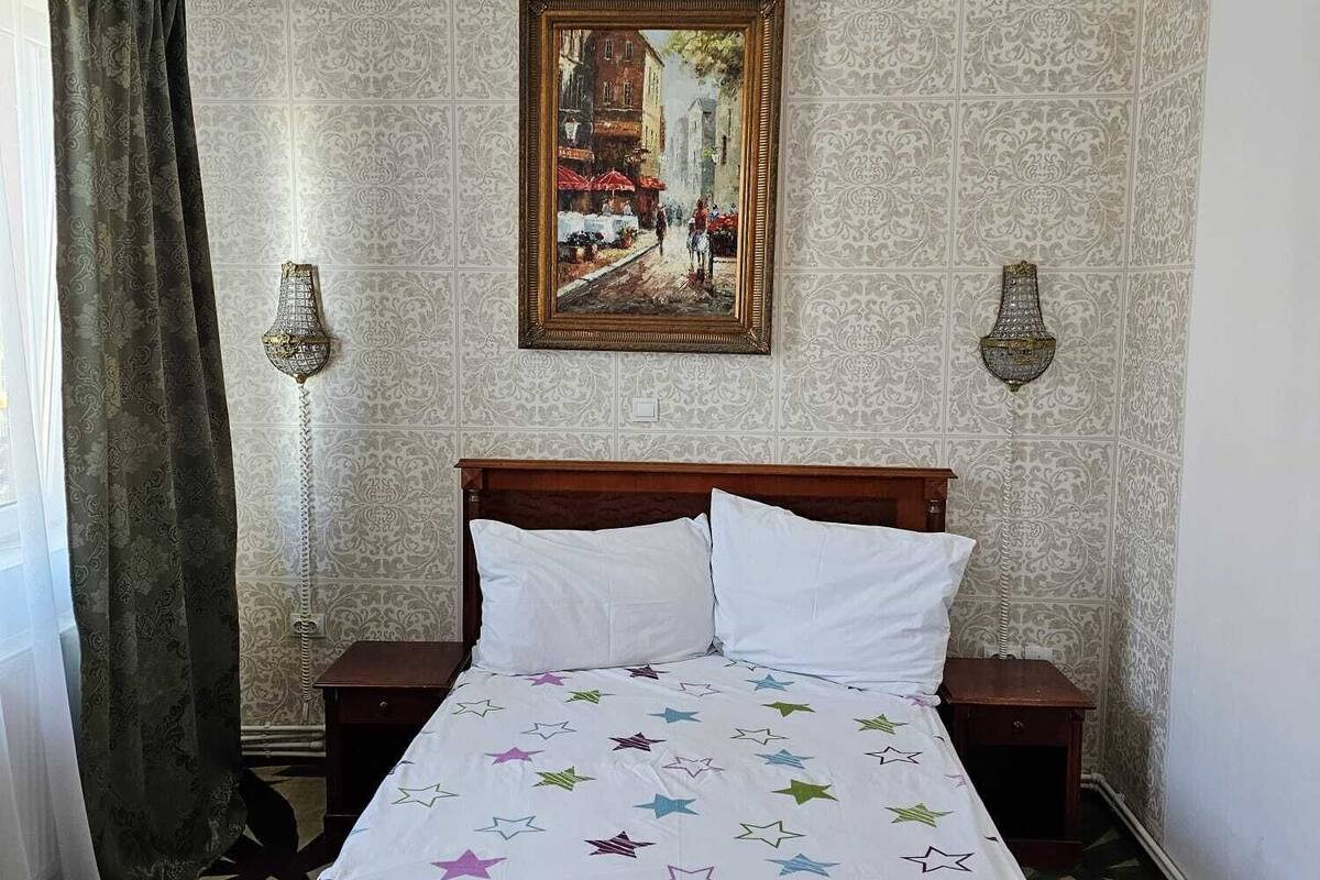 A bedroom with a bed, a painting, two wall sconces, and patterned wallpaper.