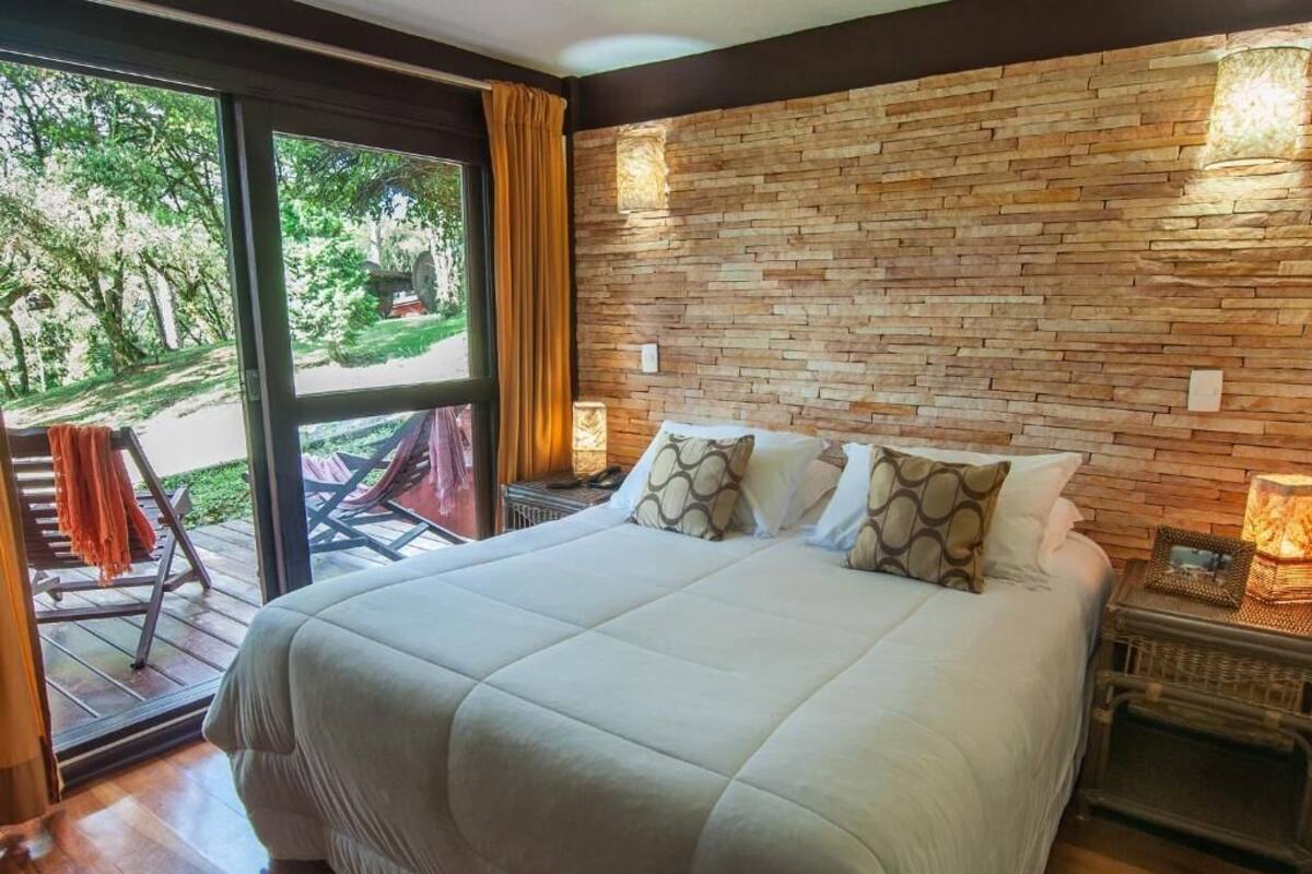 Un grand lit avec une couette blanche, deux oreillers et une tête de lit en bois. Il y a une table de chevet avec une lampe et une petite table placée à côté du lit. On aperçoit une fenêtre avec des rideaux, et une chaise est placée à l’extérieur de la fenêtre.