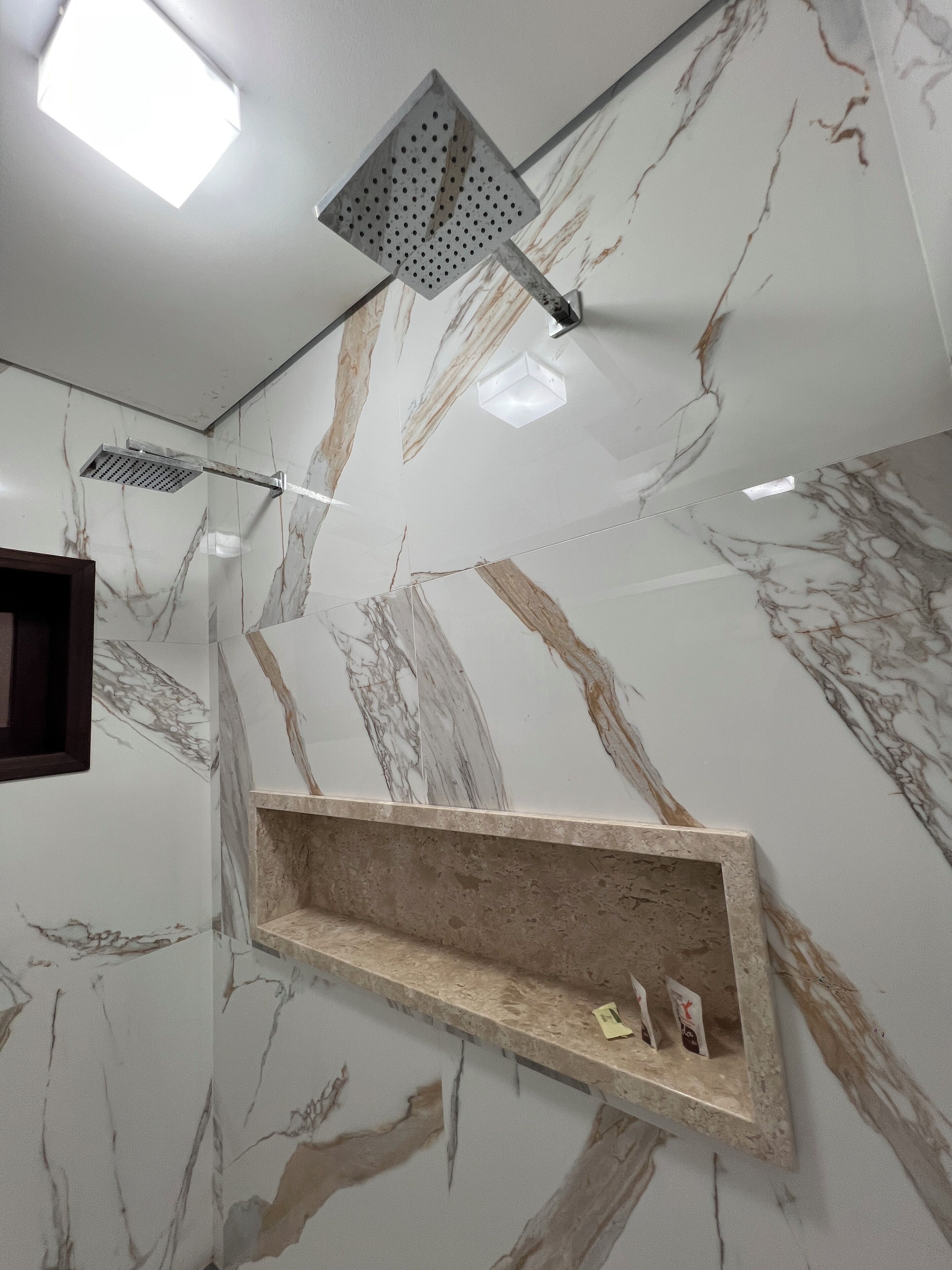 A bathroom with marble walls, a shower with a square head, and a shelf with toiletries.
