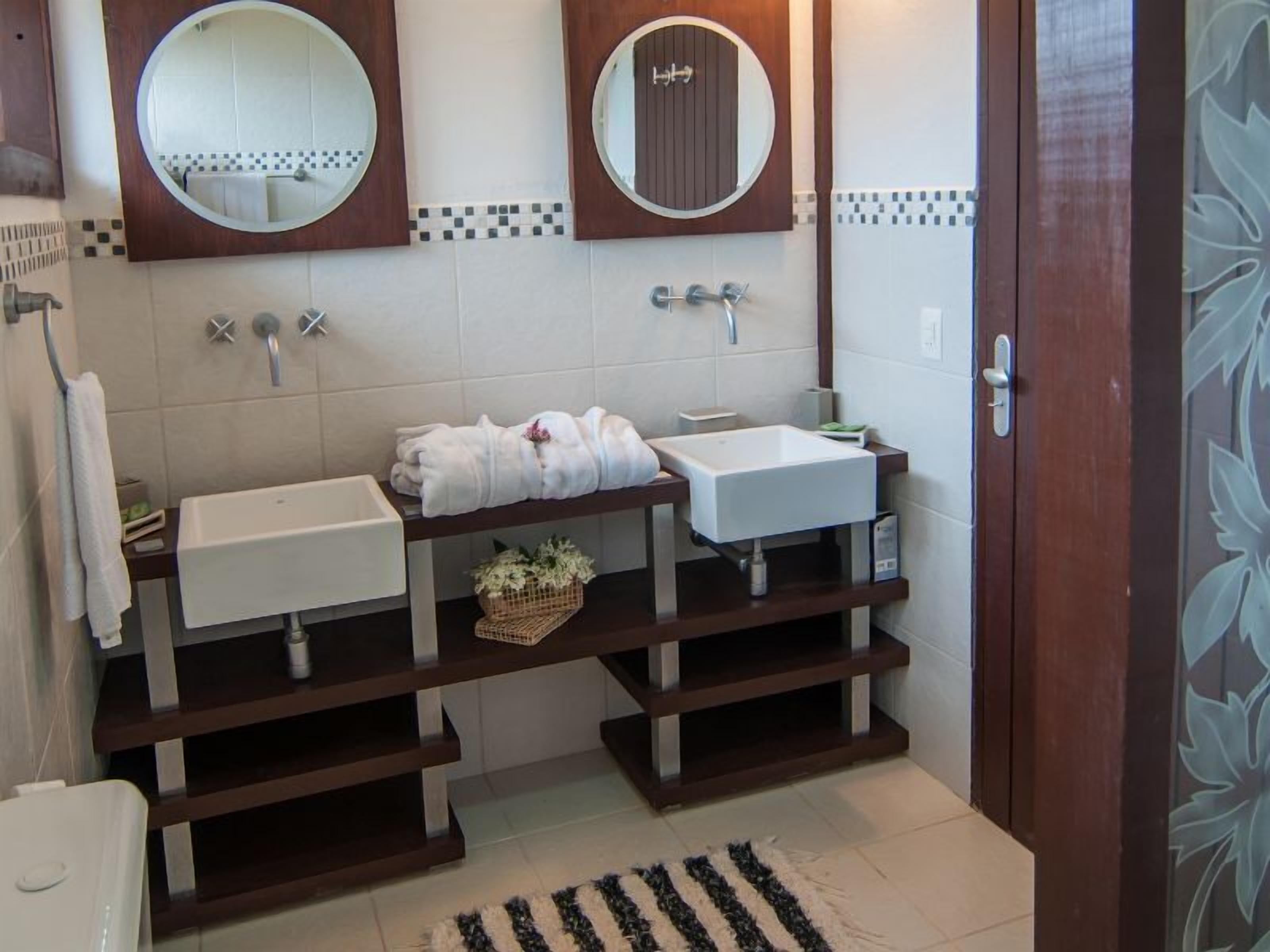Een badkamer met twee wasbakken, een toilet en een spiegel. Handdoekrek met handdoeken, een klein mandje met bloemen, en een gestreept vloerkleed.