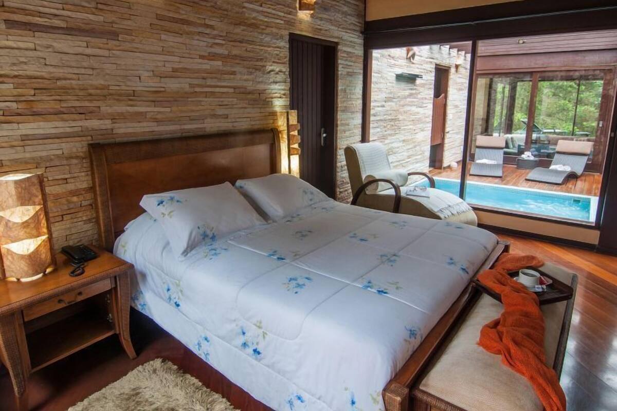 Une chambre avec un mur en pierre, un lit avec une literie à motifs floraux, une table de chevet avec une lampe, et une vue sur l’espace extérieur abritant une piscine.
