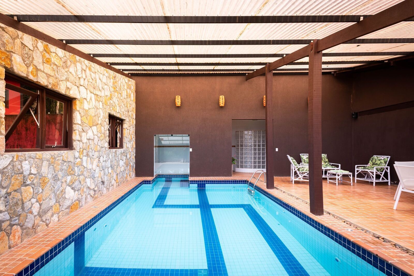 Una piscina interior con paredes de piedra y suelo de baldosas, rodeada de sillones de descanso y una zona cubierta.