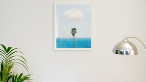 Une œuvre d’art encadrée représentant un palmier sur fond de ciel bleu et d’océan, accrochée à un mur blanc.
