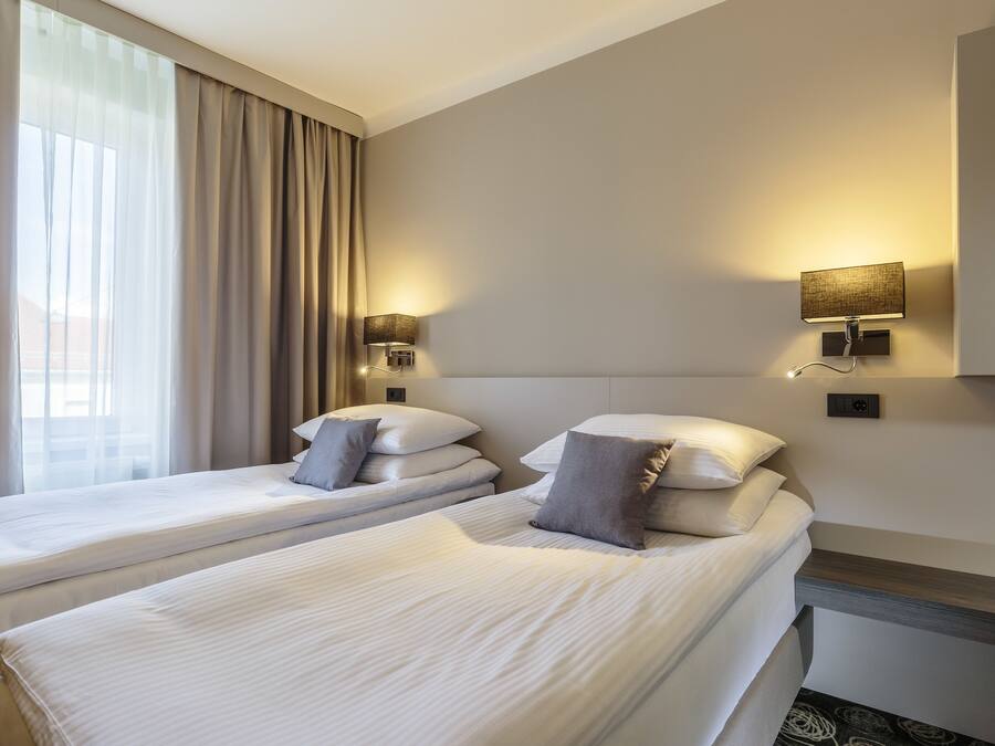 A hotel room with two beds, a large window with curtains, and wall-mounted lamps.